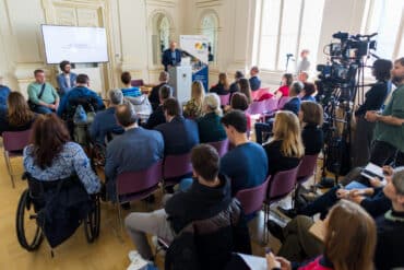 Na sliki je prostor Narodne galerije - zlata Dvorana. Za govorniškim pultom predstavlja projekt mag. Roman Rener. Pred odrom so stoli z udeleženci dogodka.