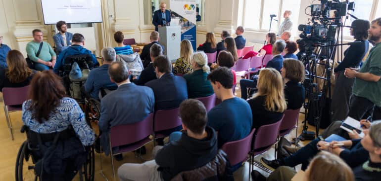 Na sliki je prostor Narodne galerije - zlata Dvorana. Za govorniškim pultom predstavlja projekt mag. Roman Rener. Pred odrom so stoli z udeleženci dogodka.